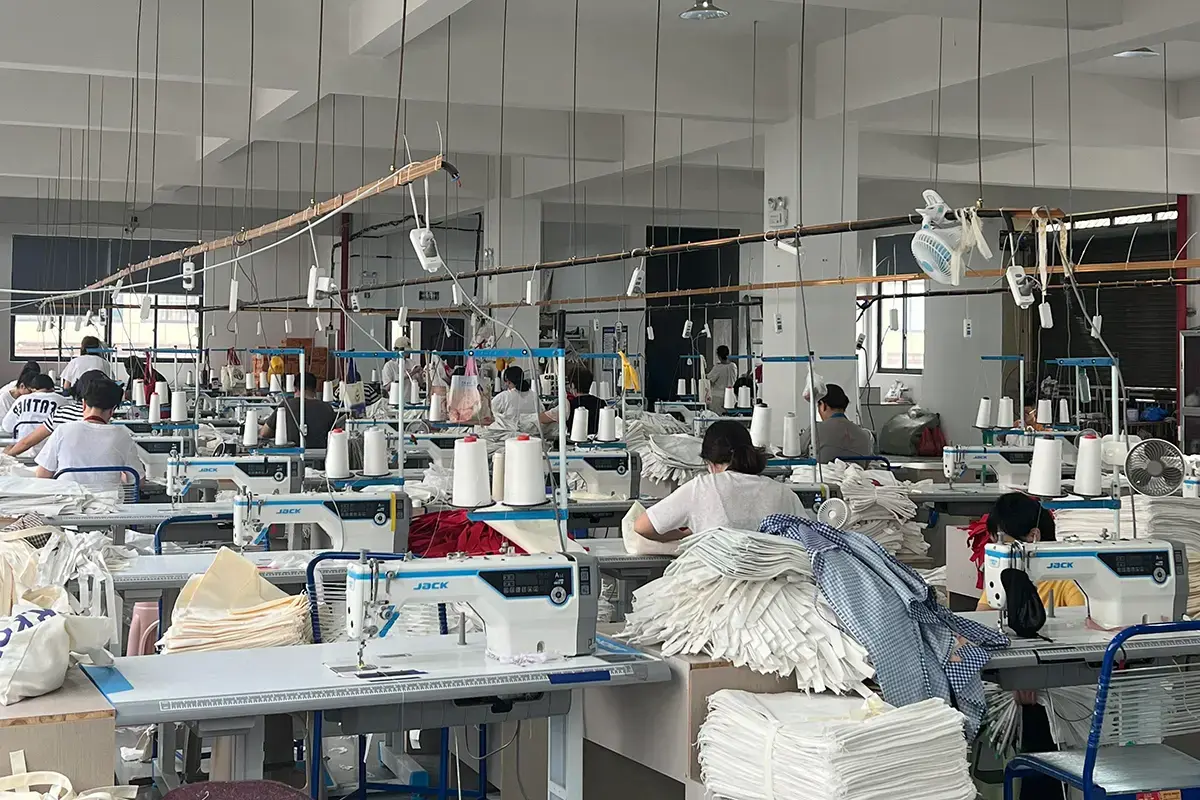 Workers sewing tote bag panels across an organized production workshop.