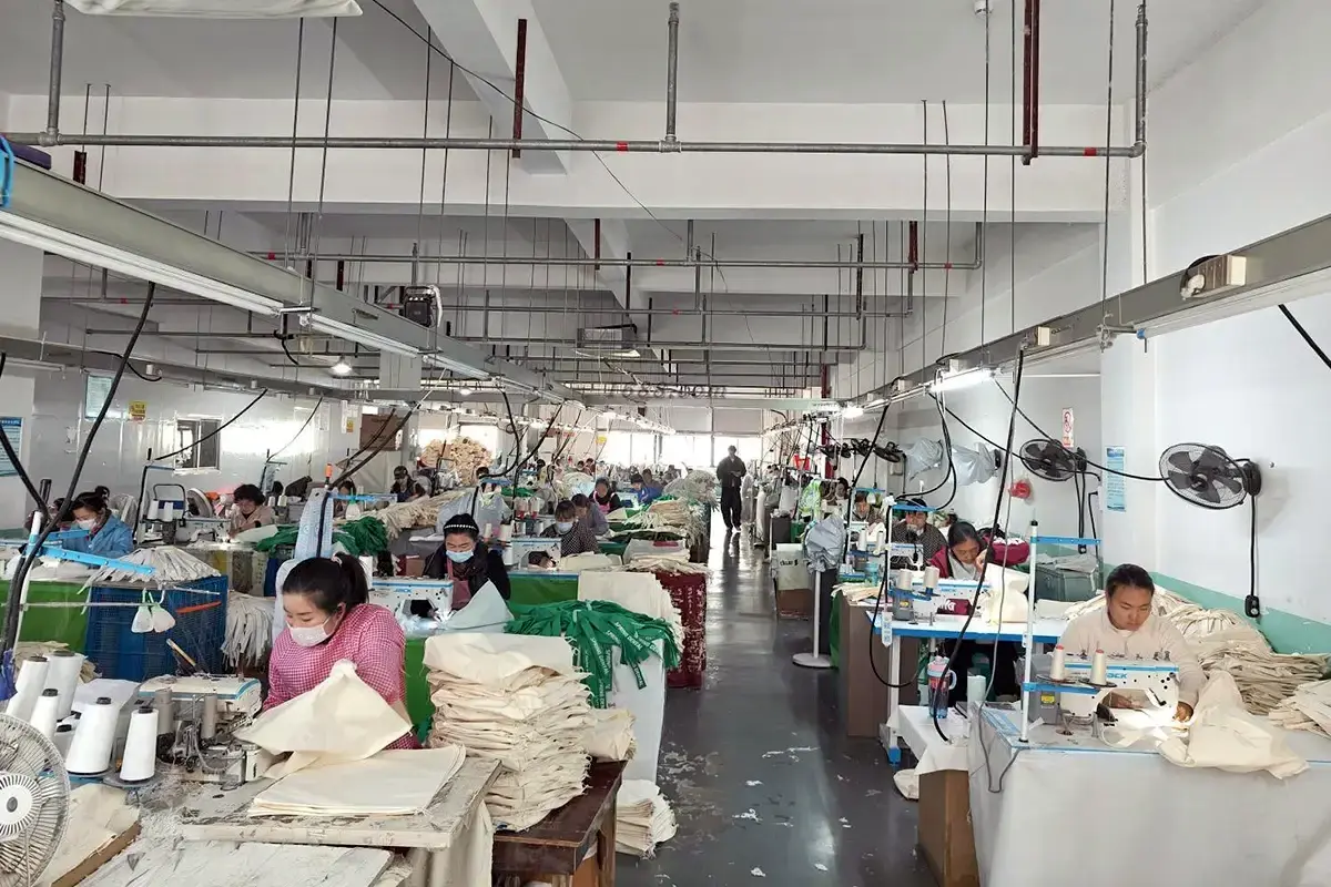 Sewing team assembling natural tote bag panels across the workshop.