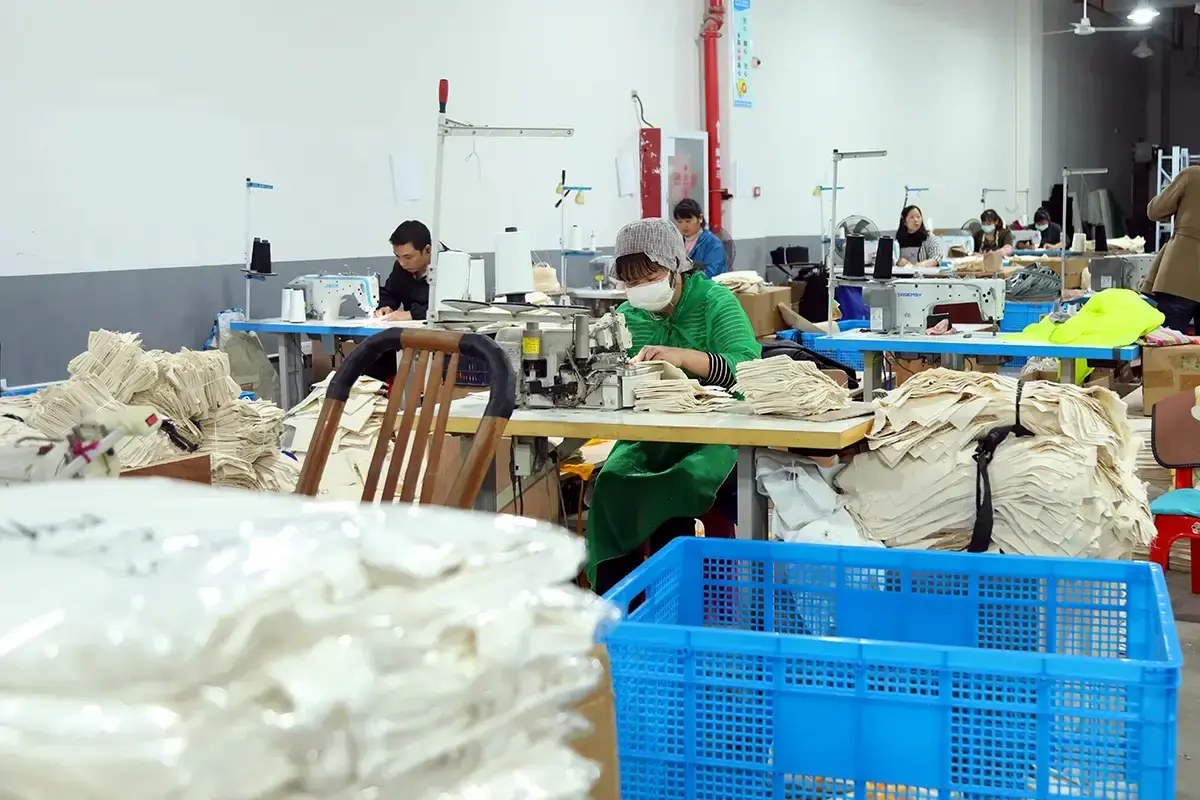Workers sewing stacks of natural canvas tote bag panels at factory stations.