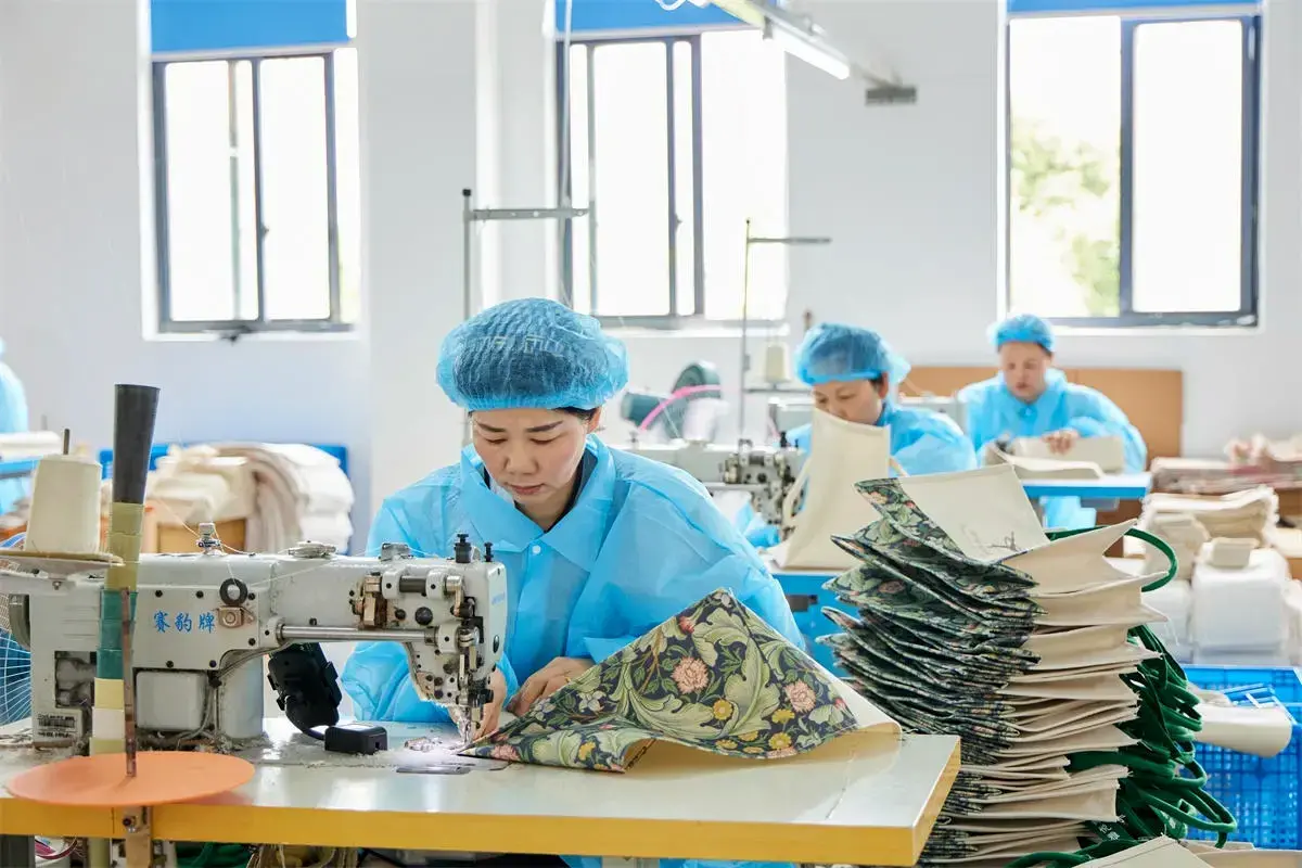 Worker sewing canvas tote panels at an industrial machine.