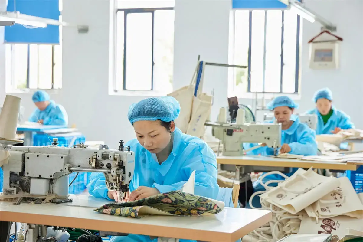 Uniformed workers sewing printed canvas tote bags at individual stations.