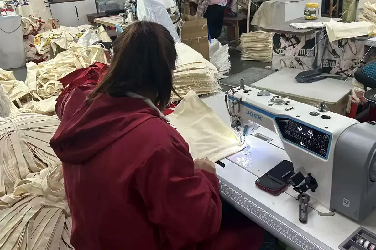 Worker sewing natural canvas tote bag panels at an industrial machine.